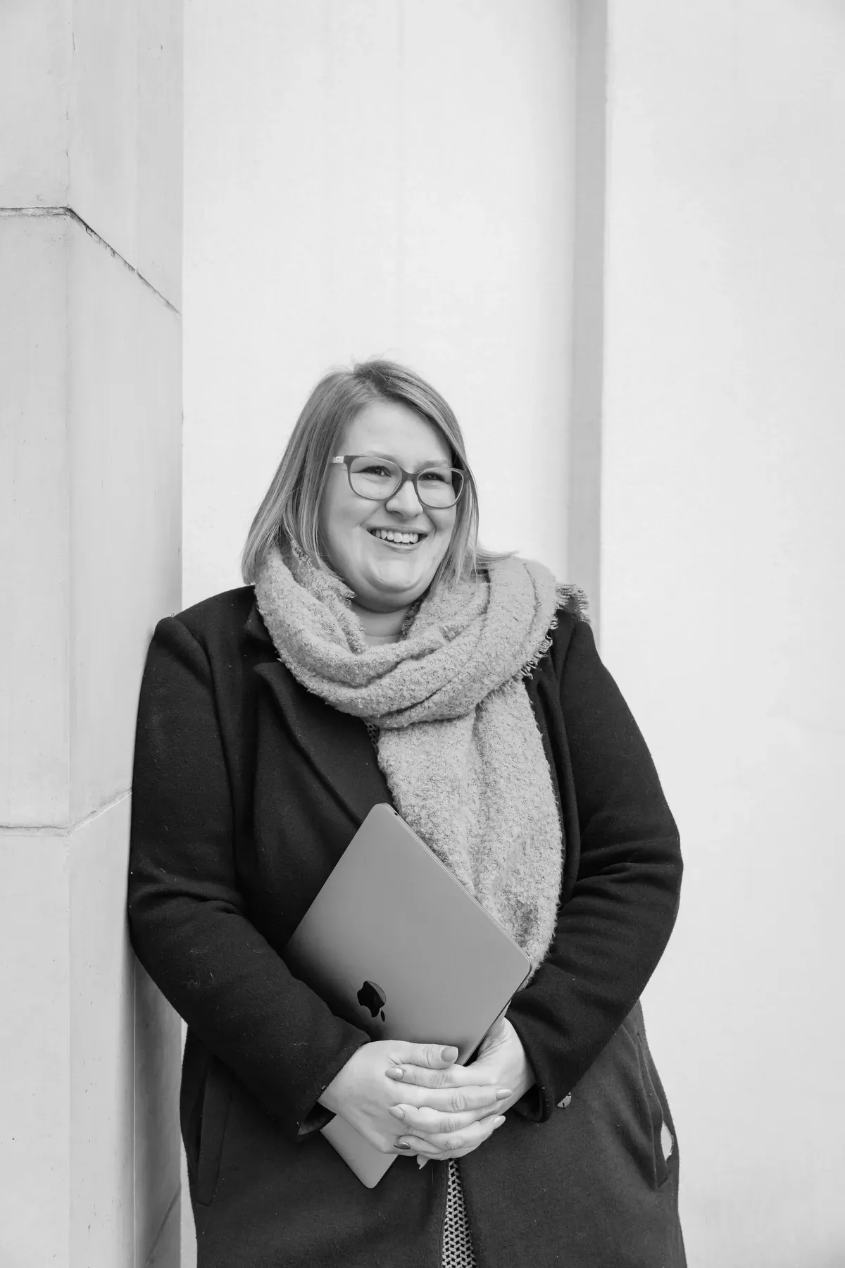 Gemma Hurlstone, smiling, holding her laptop, leaning against a pale stone building.