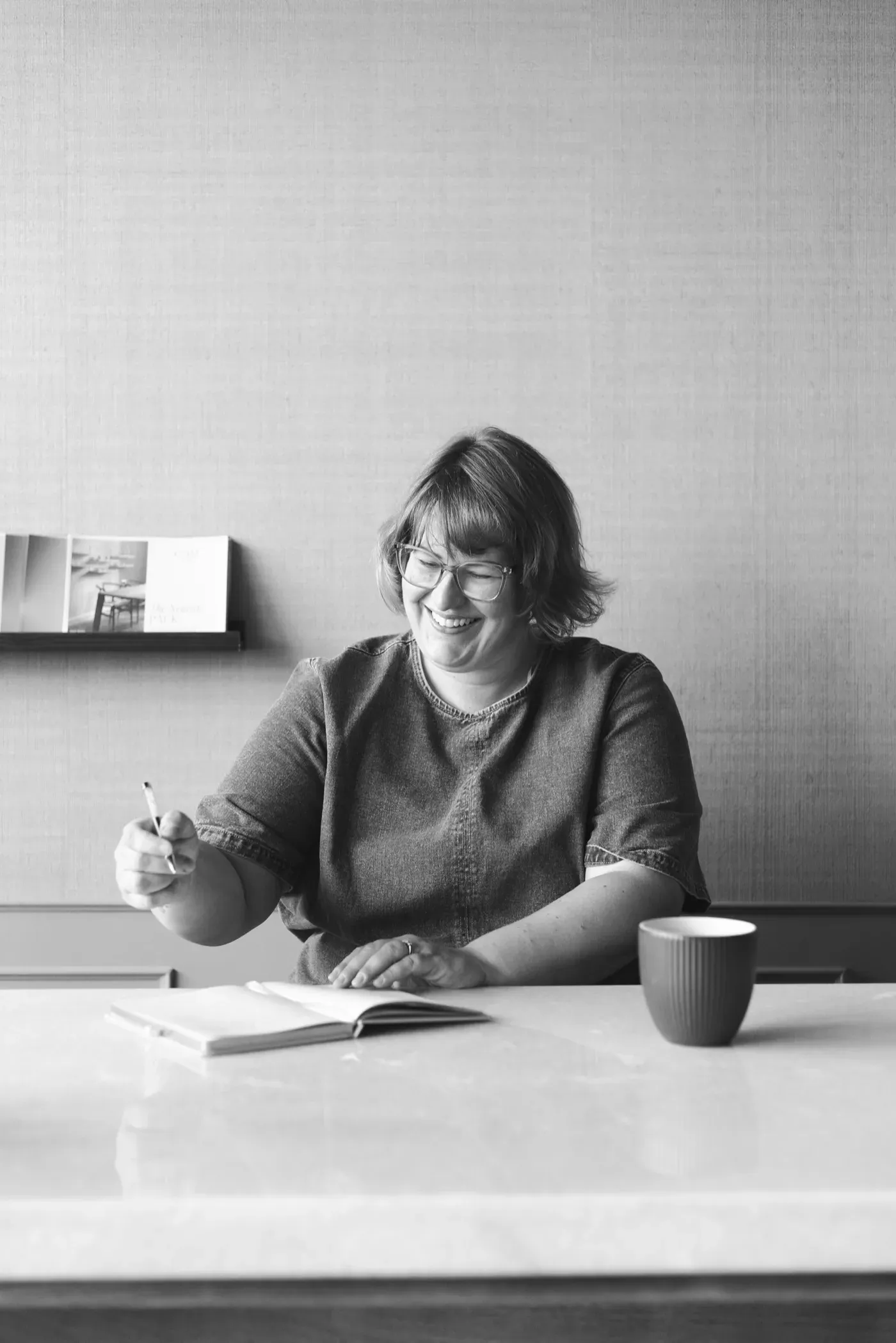 Black and white portrait of Gemma Hurlstone writing in a notebook at a table, laughing, coffee beside her.