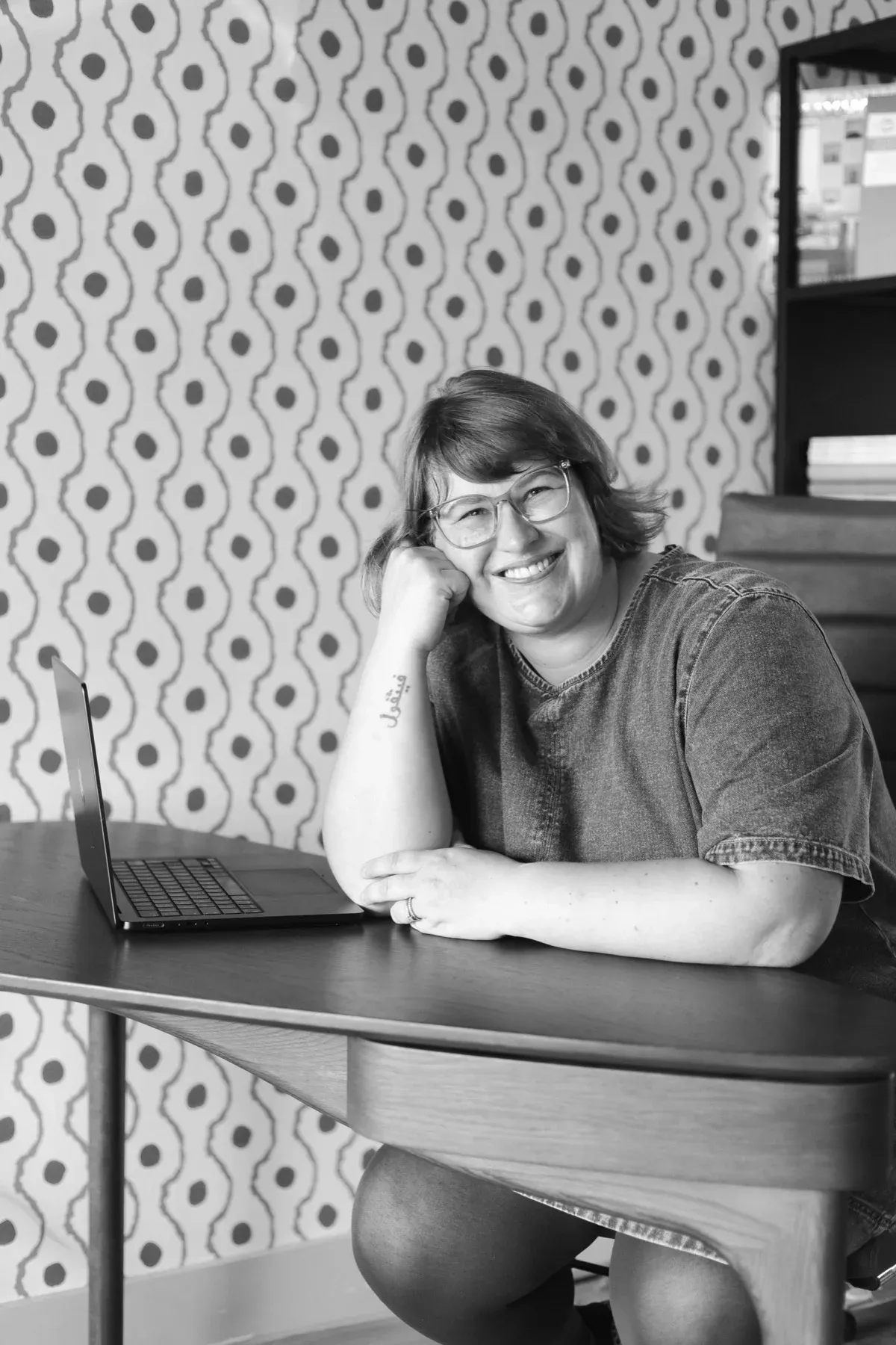 Gemma leaning on her desk and laughing, patterned wallpaper behind her.