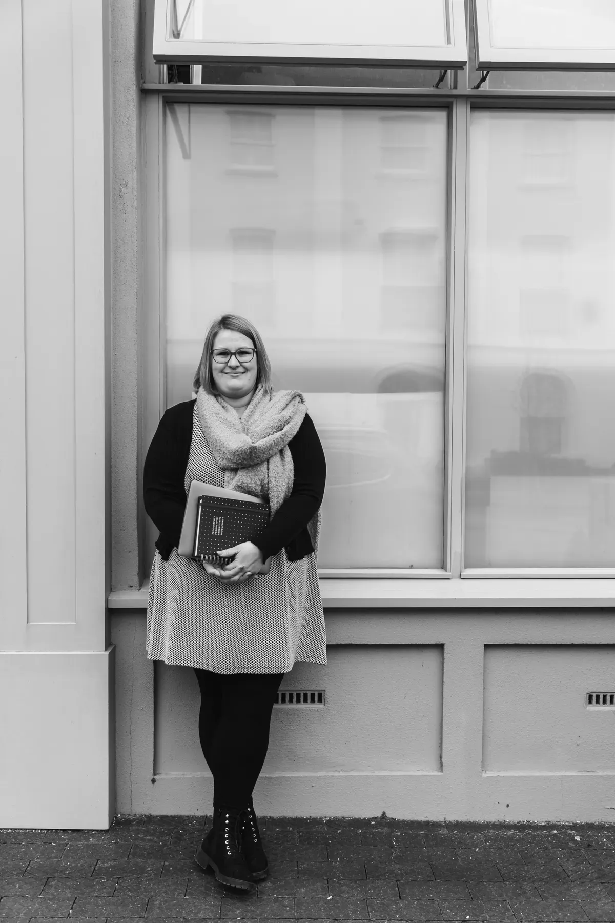 Gemma Hurlstone leaning against a shopfront window, holding notebooks, calm and grounded.