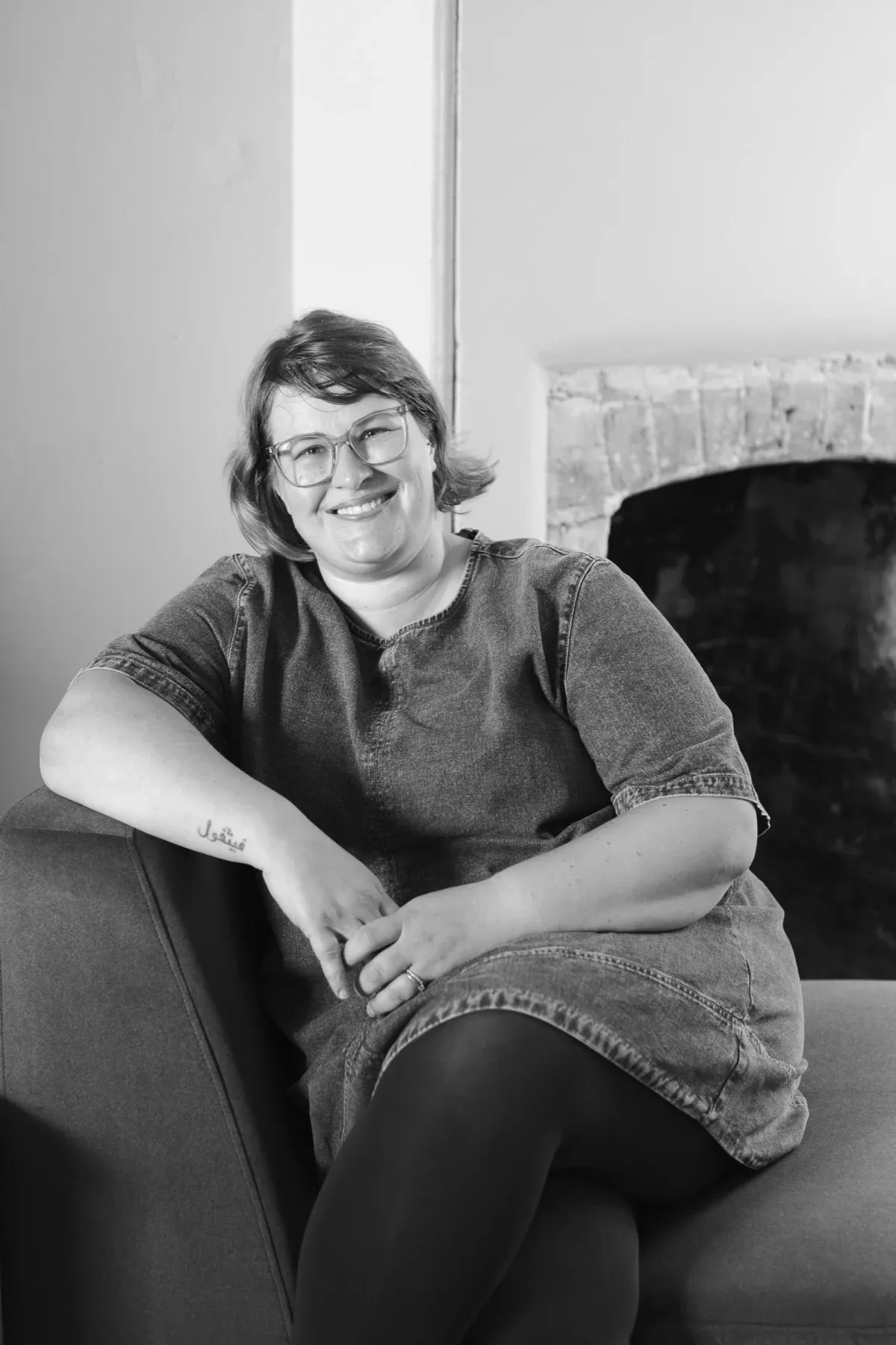 Black and white photo of Gemma Hurlstone sitting relaxed on a sofa beside an open brick fireplace, legs crossed, smiling.